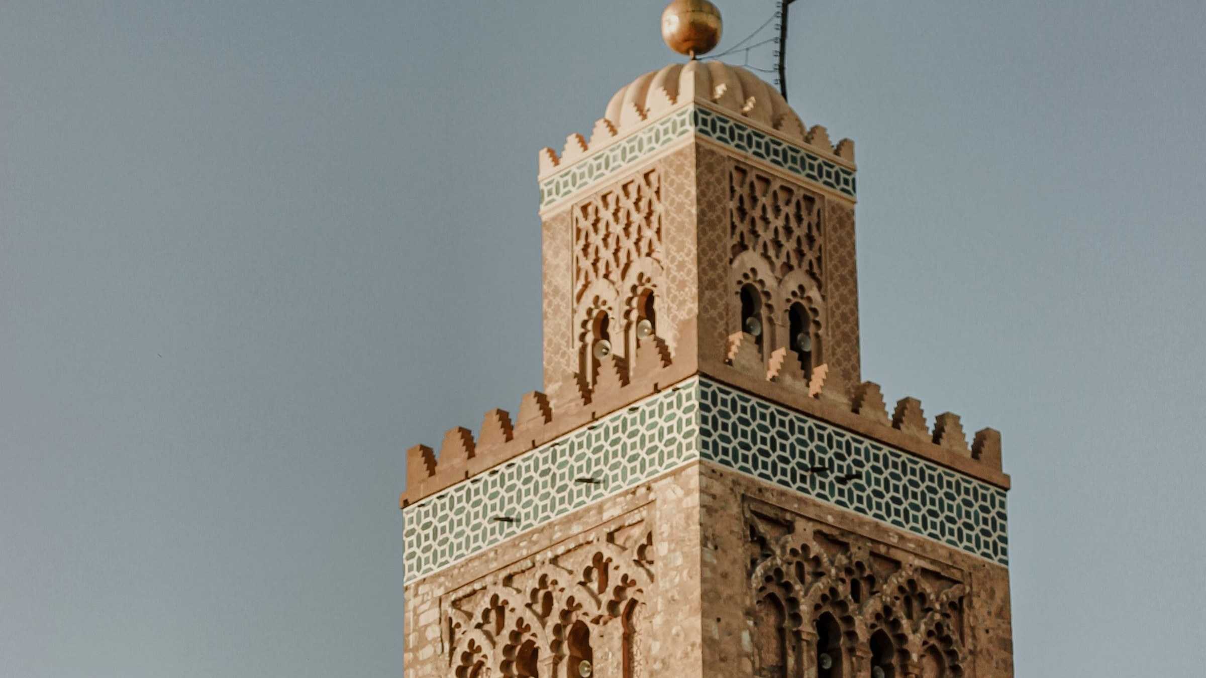 Marrakech : le sommet d’un minaret de brique ocre, décoré de carreaux turquoise, sur un ciel gris-bleu.