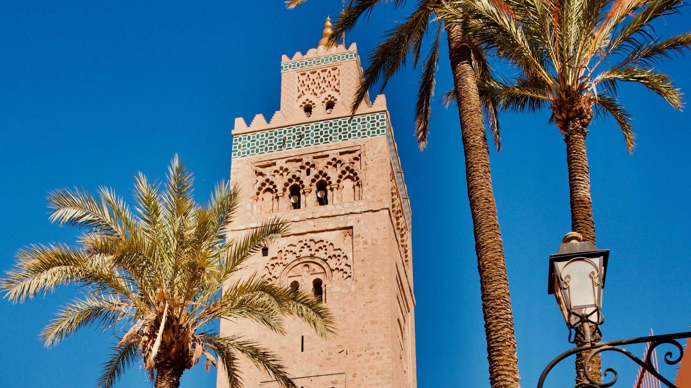Marrakech : un minaret de brique ocre domine des palmiers, au-dessus d’une porte de la médina, sous un ciel bleu.