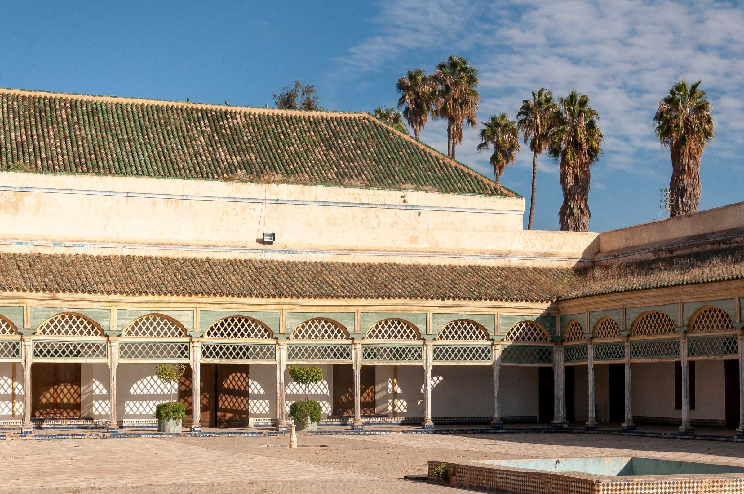Marrakech : la cour d’un ancien palais aux toits de tuiles vertes, avec des palmiers sous un ciel bleu.
